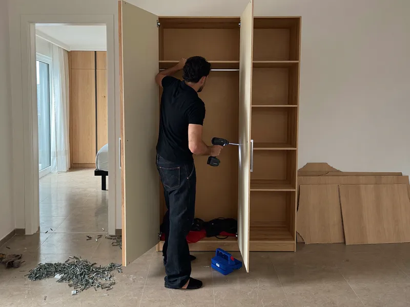 Handyman assembling a wooden wardrobe with a cordless screwdriver in an empty bedroom